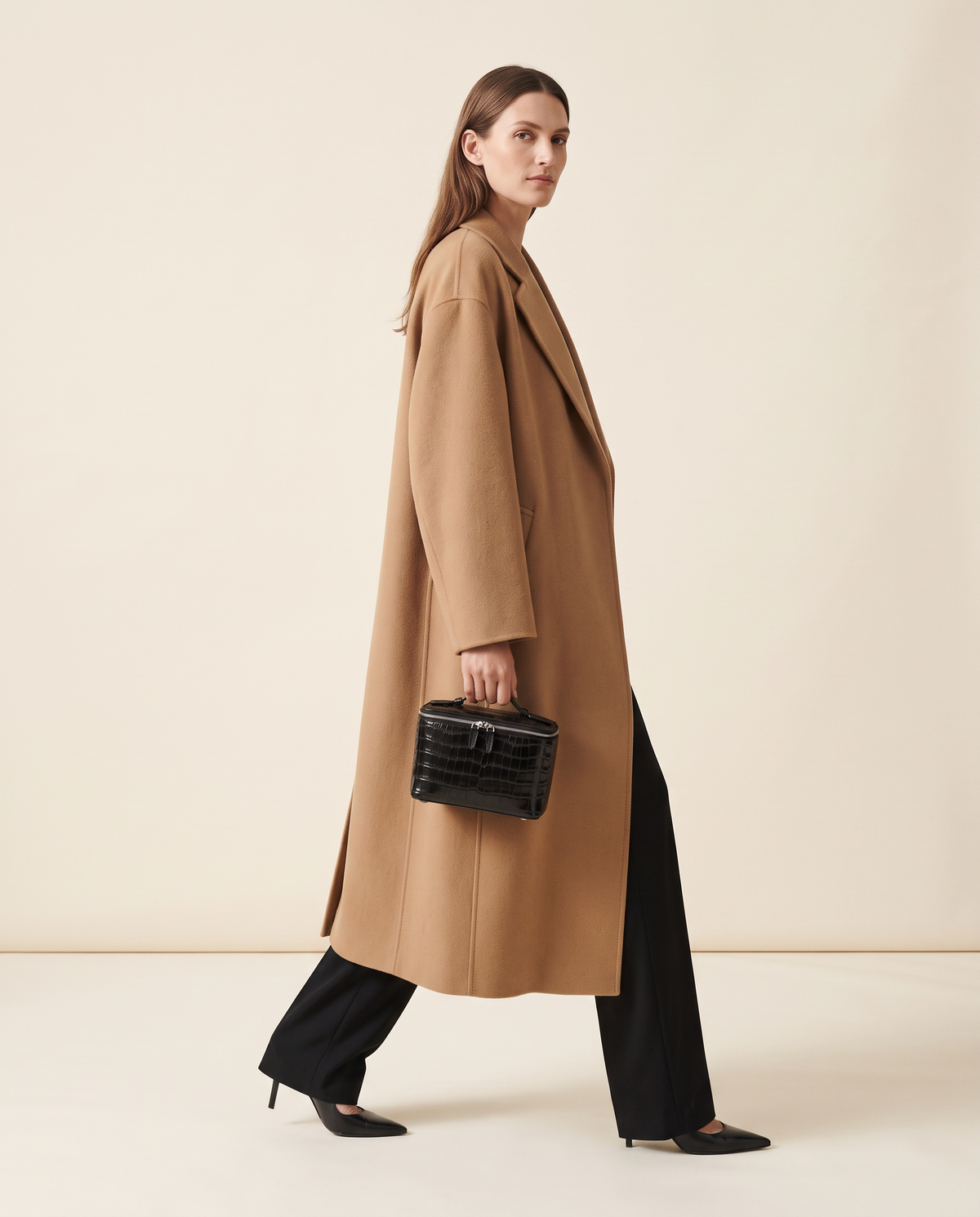 A woman in a camel coat walks carrying the small black croc Classic case